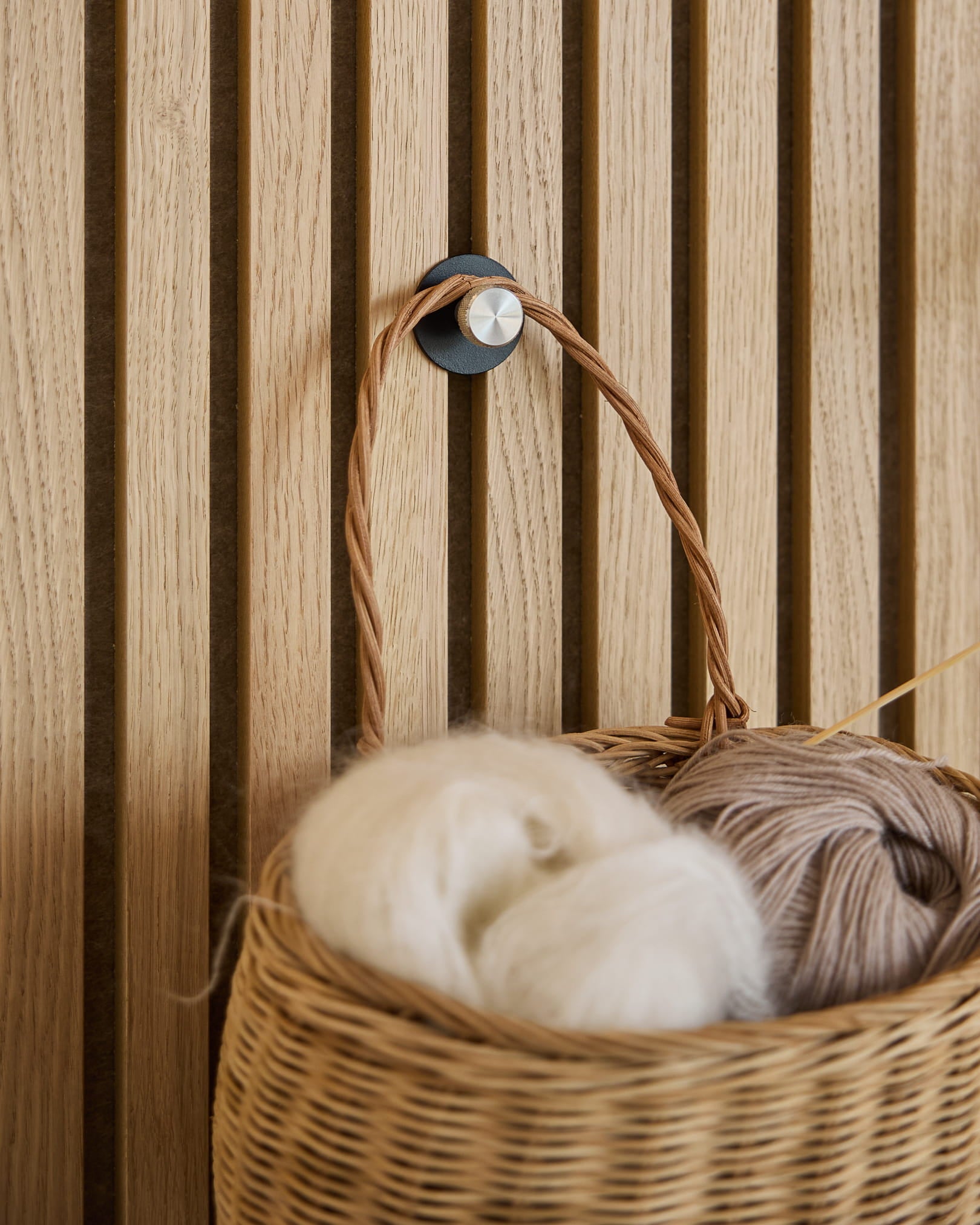Un cestino intrecciato in filato bianco e beige è appeso a un gancio appendiabiti Norr / X1, Black/Stainless Steel di WoodUpp montato su un pannello acustico a doghe Akupanel.